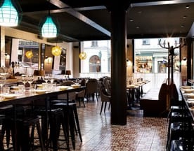 Restaurangens interiör har en modern och inbjudande design med trä- och metalldetaljer, väl upplyst med pendellampor i teal och varma toner. Det finns en mix av högryggade stolar och mjuka bänkar, samt ett långt gemensamt bord omgiven av svarta barstolar. Mönstrade plattor på golvet ger karaktär, och stora fönster släpper in naturligt ljus.