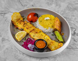 Grilled fish with a golden-brown exterior, served with saffron-infused rice, grilled tomato, chargrilled green vegetable, lemon wedge, edible flowers, and a small bowl of spicy or tangy sauce.