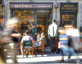 Fachada encantadora do restaurante "Botánico" com grandes janelas, área de estar exterior com cadeiras de madeira e um ambiente animado, ideal para refeições e bebidas.