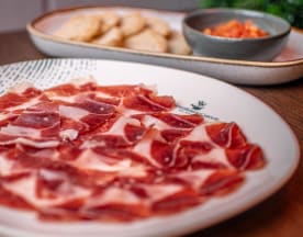 Plato de jamón curado en rodajas finas, con un color marrón rico y vetas de grasa blanca, acompañado de una crema para untar y galletas redondas.