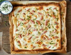 Tarte flambée avec une croûte dorée et croustillante, garnie d'oignons tranchés et de morceaux de jambon, accompagnée de ciboulette et d'une sauce crémeuse.