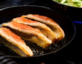 Três filés de salmão a grelhar numa frigideira de ferro fundido, com crosta dourada e carne húmida e flocosa, acompanhados por legumes salteados.
