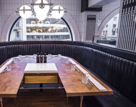 Interior moderno de restaurante con un amplio sofá curvo de cuero oscuro, mesa de madera rica y diseño único, iluminado por lámparas colgantes elegantes.