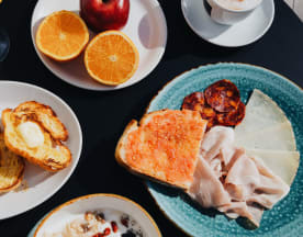 Desayuno variado con tostadas con mantequilla y mermelada, pan con tomate y embutido, yogur cremoso con bayas de goji, y frutas frescas como naranjas y manzana.