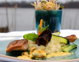 Das Gericht besteht aus einem perfekt gebratenen Fischfilet mit knuspriger Haut, serviert mit cremigem Risotto, das mit Meeresfrüchten, frischen grünen Gemüse wie zartem Spargel und dünn geschnittenem Zucchini verfeinert ist. Eine zarte Garnitur aus jungen Kräutern und einer kleinen Karottenlocke rundet die Präsentation ab.