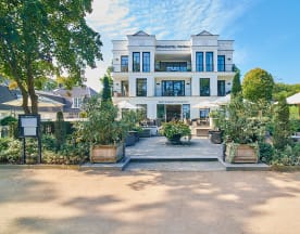 Modernes Restaurant mit weißer Fassade, großen Glasfenstern, mehreren Ebenen und einer großzügigen Terrasse mit Sonnenschirmen. Um den Eingang herum befinden sich gepflegte Pflanzenkübel, die die einladende Atmosphäre unterstreichen.