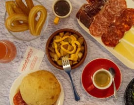 Assortiment de plats comprenant du pain doré, des tranches de charcuterie, des wedges de fromage crémeux, des bananes plantains frites, du café noir, des churros sucrés et un verre de jus d'orange.