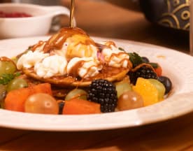 Une pile de pancakes moelleux arrosés de sirop brillant, garnis d'une généreuse cuillerée de crème fouettée et entourés d'un assortiment de fruits frais, dont des mûres, des raisins et des segments d'orange.