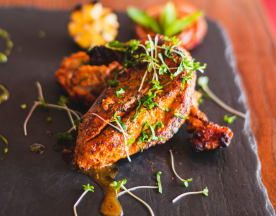 Grilled chicken with a golden-brown crust, garnished with microgreens, served with chargrilled corn, roasted tomato, and drizzled with herb-infused oil and green sauce.