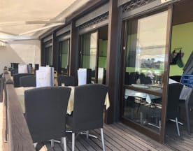 Área de refeições ao ar livre com mesas cobertas por toalhas claras e cadeiras de vime escuras, sob uma cobertura que proporciona sombra e conforto. Grandes janelas de vidro oferecem vista para um interior bem iluminado, criando um ambiente acolhedor e convidativo.