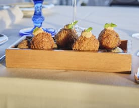 Croquetes dourados e crocantes, moldados em pequenas bolas, cobertos com um molho cremoso e guarnecidos com ervas frescas.
