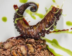 Tentáculo de polvo grelhado com exterior carbonizado, servido sobre um risotto cremoso de frutos do mar, guarnecido com microverdes e molhos coloridos.