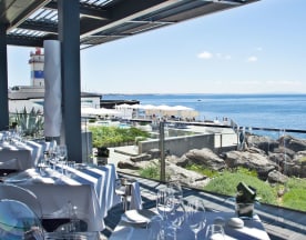 Área de refeições ao ar livre com vista para o oceano, mesas elegantemente decoradas, céu azul e águas calmas, criando uma atmosfera tranquila e relaxante.