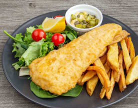 Un morceau de poisson doré et croustillant, probablement du cabillaud, accompagné de frites épaisses, d'une salade de légumes frais, de tomates cerises et d'un quartier de citron, ainsi qu'un petit bol de cornichons.