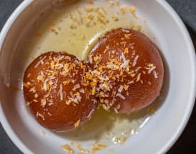 Dois doces redondos, provavelmente gulab jamun, mergulhados em xarope, com exterior brilhante e interior macio, cobertos com coco ralado.
