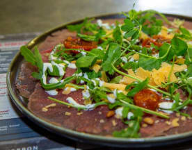 Dunne plakjes gezouten vlees op een schaal, gegarneerd met verse rucola en microgroenten, zongedroogde tomaten, een romige saus, geroosterde pijnboompitten en geschaafde kaas voor een kleurrijke en smaakvolle presentatie.