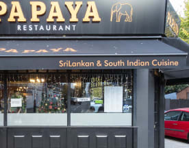 The exterior of Papaya Restaurant features a dark facade with golden lettering, large windows showcasing festive decorations, and an awning with string lights, creating a warm and inviting atmosphere for Sri Lankan and South Indian cuisine.