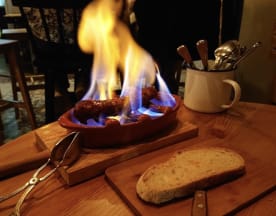 Salsichas flambadas, apresentando um espetáculo de chamas azuis, acompanhadas por uma fatia de pão rústico ideal para absorver os sucos saborosos.