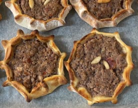 Uma seleção de pequenas tortas redondas com uma crosta rústica e dourada. Cada torta tem uma borda ligeiramente elevada, assemelhando-se a uma flor. O recheio é uma mistura de carne picada, possivelmente de cordeiro ou vaca, temperada com especiarias e ervas, e coberta com pinhões inteiros para adicionar textura. Uma opção saborosa e robusta, ideal para partilhar.