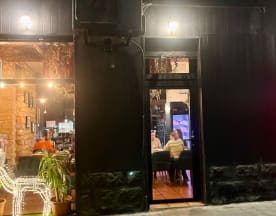 Exterior de restaurante moderno con fachada negra, luces cálidas en el interior y ambiente acogedor.