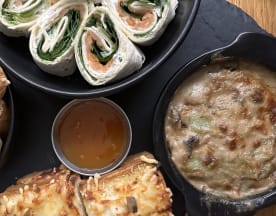 Variété de plats disposés sur une surface en ardoise sombre, comprenant un gratin crémeux de légumes et champignons, des wraps roulés avec des légumes frais et du saumon fumé, des boules dorées croustillantes, accompagnées d'une petite sauce sucrée, et des morceaux de pain plat grillé avec du fromage fondu et des légumes.
