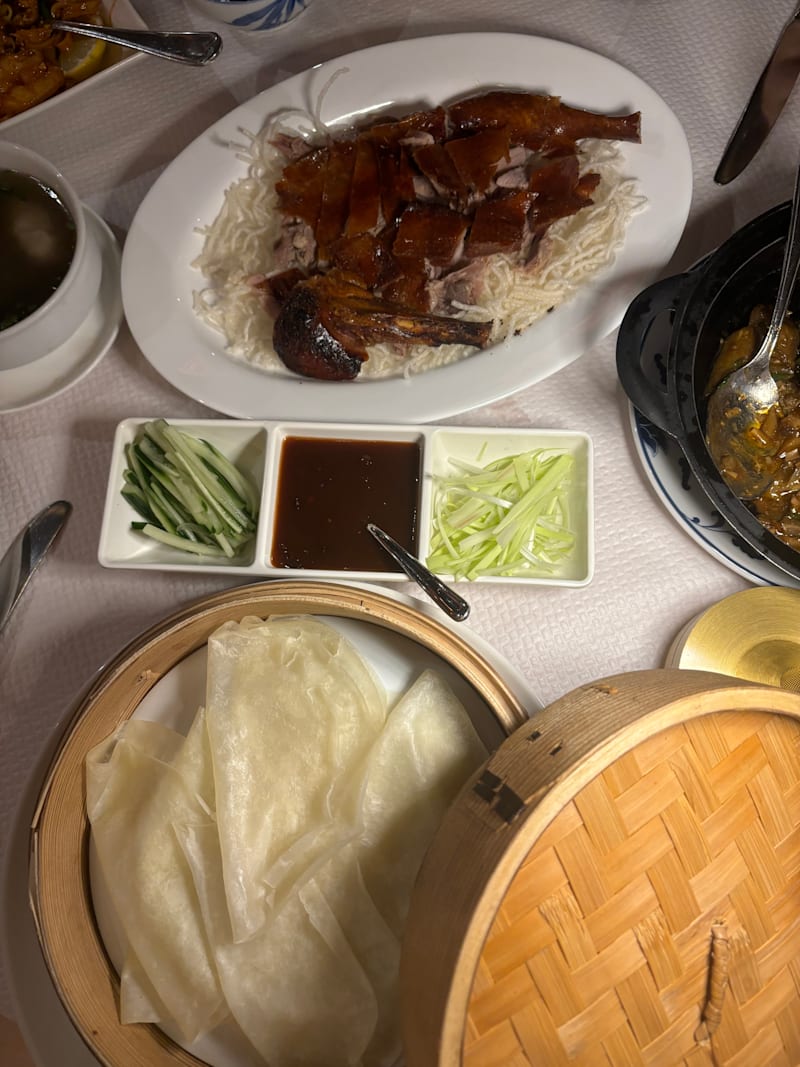 Sélection de plats avec un canard rôti, servi sur un lit de chou finement émincé, accompagné de sauces pour tremper, de wrappers de riz translucides, de concombre et d'oignons verts, ainsi qu'un bol de soupe.