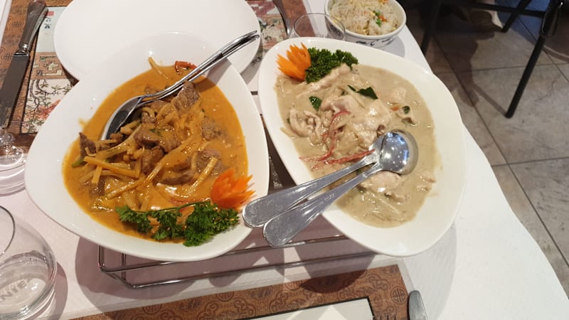 Deux plats distincts : à gauche, un curry épais avec des morceaux tendres de viande dans une sauce crémeuse orange, garni de pousses de bambou et d'herbes fraîches. À droite, un plat léger à base de noix de coco avec des morceaux de poulet ou de fruits de mer dans une sauce crémeuse pâle, décoré de feuilles de basilic et de quelques tranches de piment rouge. Les deux plats sont accompagnés d'un bol de riz moelleux.