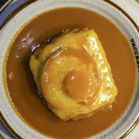 Pão em forma quadrada, possivelmente um pudim de pão salgado, coberto com uma gema de ovo brilhante e acompanhada de um molho laranja profundo, criando uma refeição reconfortante e indulgente.