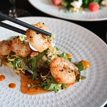 Gegrilde coquilles geserveerd op een bed van gemengde groenten met een rijke, pittige saus en een topping van gemalen noten voor extra crunch.