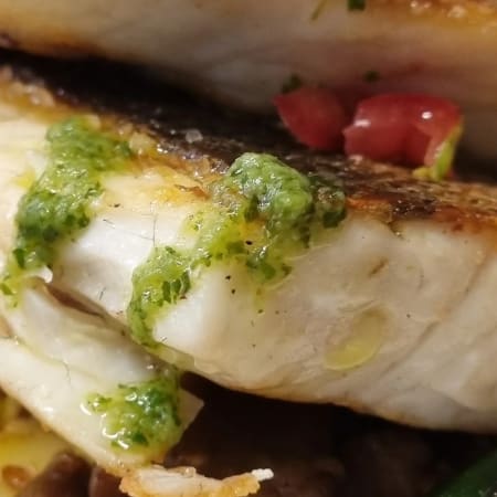 Perfekt gebratenes Fischfilet mit goldbrauner Kruste, serviert mit einer grünen Kräutersauce, sautierten Gemüse wie Tomaten und Paprika sowie knusprigen, gerösteten Kartoffeln.