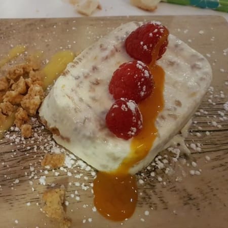 Cremiges Dessert auf einem Holzbrett, bestehend aus einem rechteckigen Stück Schaumcreme, garniert mit Himbeeren und einem Fruchtcoulis, umgeben von zerbröselten Keksen und Puderzucker.