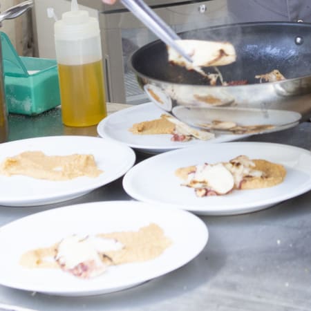 Der Koch richtet ein Gericht an, das eine cremige Sauce als Basis hat, und fügt ein perfekt gebratenes Stück Fisch oder Fleisch hinzu. Ein grobkörniges Topping, möglicherweise aus Semmelbröseln oder Nüssen, wird zur Garnierung verwendet.