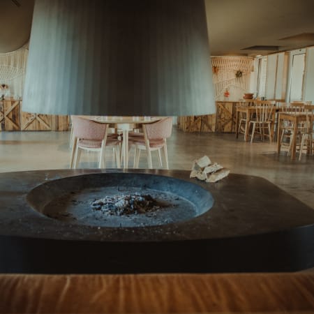 Interior de restaurante moderno com lareira circular central, rodeada por mesas e cadeiras de madeira, paredes claras e grandes janelas que permitem a entrada de luz natural, criando uma atmosfera acolhedora.