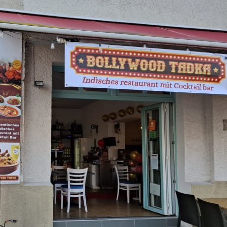 Das Restaurant "Bollywood Tadka" präsentiert eine lebhafte Atmosphäre mit bunten roten und gelben Ballons am Eingang. Ein großes Banner hebt die indische Küche und die Cocktailbar hervor. Die Fenster bieten einen einladenden Blick auf den Innenraum mit weißen Stühlen und einer gemütlichen Einrichtung, was auf einen einladenden Ort für den Genuss von aromatischen indischen Gerichten und Getränken hinweist.