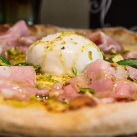 Pizza recién horneada con corteza delgada, jamón, burrata cremosa, pistachos triturados y albahaca fresca.