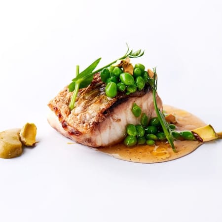 Pescado a la parrilla con guisantes frescos, hierbas delicadas y salsa cremosa, decorado con nueces enteras.
