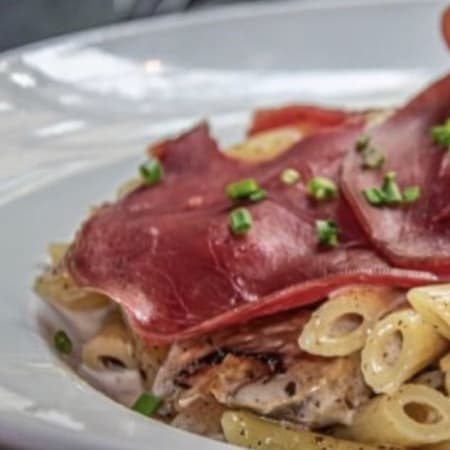 Plat copieux de pâtes, probablement des penne, surmontées de tranches de viande séchée, accompagnées d'une sauce légère et garnies d'herbes vertes finement hachées.
