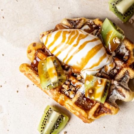 Gaufre dorée surmontée d'une boule de glace à la vanille et nappée de sauce caramel, accompagnée de tranches de kiwi frais et de noix hachées, le tout saupoudré de sucre glace.