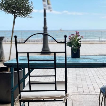 Configuración de comedor al aire libre con una mesa rústica azul cerca de la playa, decorada con flores frescas y rodeada de sillas, todo en un ambiente relajante junto al mar.