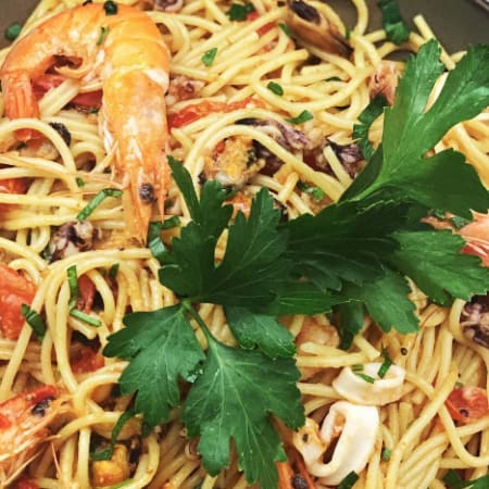 Portion généreuse de spaghetti avec de grosses crevettes et du calamar tendre, enrobée d'une sauce dorée à l'ail et aux herbes, parsemée de tomates fraîches et garnie de persil.