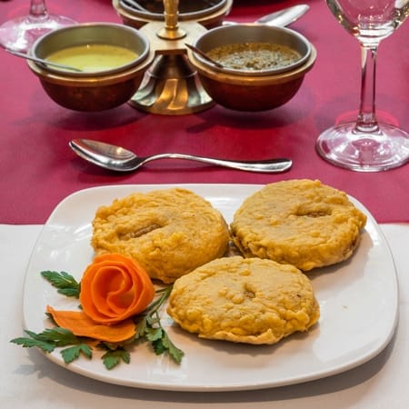 Tres croquetas doradas y crujientes, probablemente de lentejas o garbanzos, acompañadas de salsas verde y amarilla, y decoradas con una rosa de zanahoria y hierbas frescas.