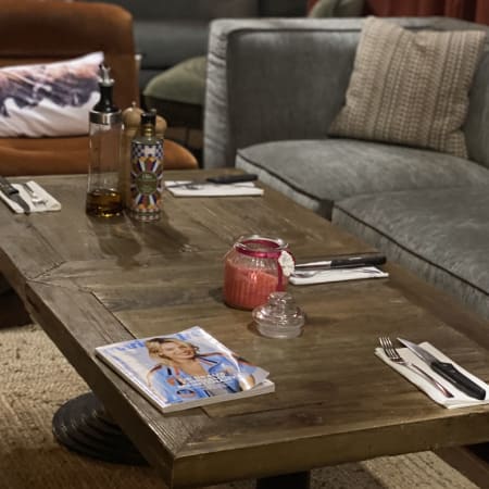Espace de restauration chaleureux avec une table en bois rustique, des bouteilles en verre, une bougie décorative et un canapé gris confortable, créant une ambiance intime et décontractée.