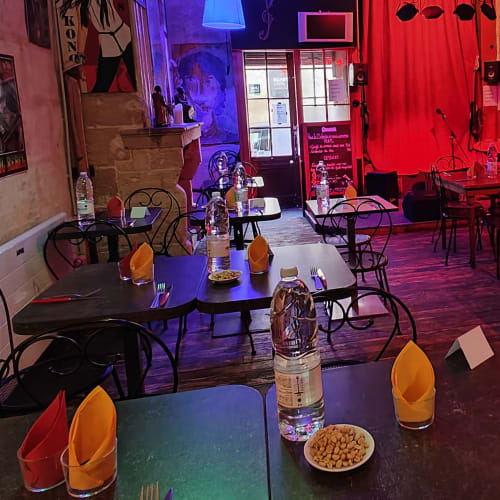 Intérieur de restaurant chaleureux avec des tables en bois foncé, nappes jaunes élégamment pliées, œuvres d'art colorées aux murs et éclairage doux en bleu et rouge, créant une ambiance vivante et détendue.