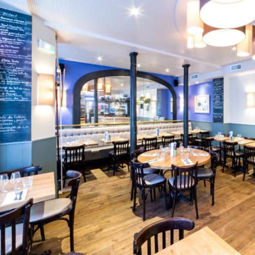 Intérieur chaleureux avec un sol en bois, des tables et chaises en bois, des murs aux tons doux, des éléments décoratifs comme des plantes et des tableaux, et un éclairage doux créant une ambiance conviviale.