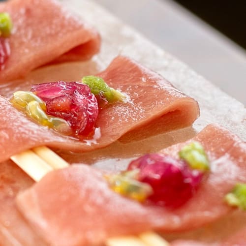 Fatias delicadas de peixe cru, provavelmente atum, dispostas sobre um bloco de sal, cobertas com pequenas porções de um condimento verde brilhante e guarnecidas com pedaços de cebola roxa ou rábano.