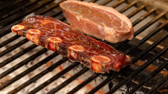 Uma seleção de carnes grelhadas sobre chama aberta, destacando um corte grosso de costela de vaca com um glaze escuro e rico, e um corte menor, possivelmente de porco ou vaca, com uma cor mais clara e marmoreio. As marcas da grelha indicam um cozimento perfeito, com sucos a realçar o sabor.