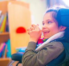 A boy in a wheelchair is smiling to someone off camera, he is clasping his hands in front of his face.