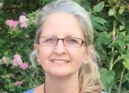 A white lady with blonde and grey curly hair and wearing glasses smiles at the camera