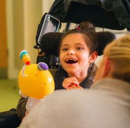 A child in a wheelchair smiles. They are looking at a brightly coloured toy, which is being show to them by a carer.