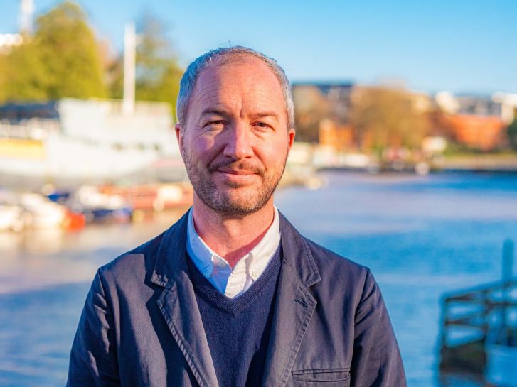 A man smiles at the camera. He is wearing a blue suit. Behind him is Bristol harbourside
